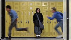 a woman standing in front of yellow lockers with her legs spread out and two men running behind her