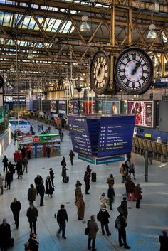 many people are walking around in an indoor area with large clocks hanging from the ceiling