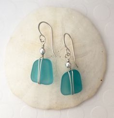 two sea glass pieces are hanging from silver earwires on a white stone slab