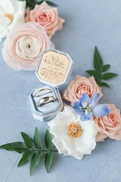 two wedding rings in an open box surrounded by pink and white flowers on a blue background