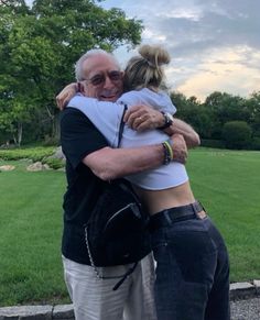 an older man and woman hug each other in front of a grassy area with trees