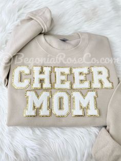 a sweatshirt with the word cheer mom in gold sequins sitting on a furry surface