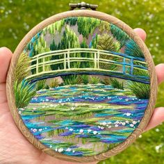 a hand holding up a small embroidery art piece with water lilies and a bridge in the background