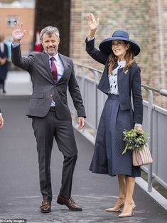 two people in suits and hats wave at the camera while standing on a bridge with other people