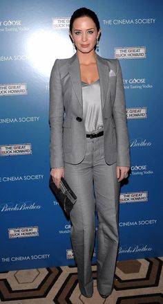 a woman standing in front of a blue wall wearing a gray suit and black purse