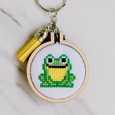 a cross stitch frog keychain with a yellow tassel hanging from it's side