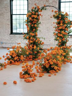 an arrangement of orange flowers and greenery in front of a brick wall with arched windows