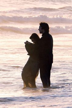 a man and woman standing in the ocean at sunset