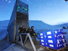 two men are standing outside in the snow near some crates and an object that looks like a tower