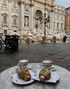 two coffee cups and pastries sit on a table in front of a fountain