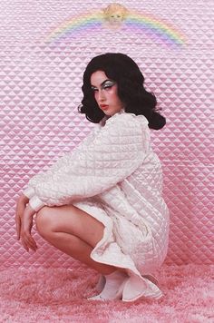 a woman kneeling down in front of a pink wall with a rainbow above her head