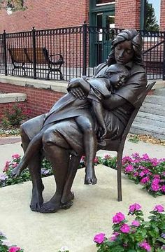 a statue of a woman sitting on a chair in front of some flowers and bushes