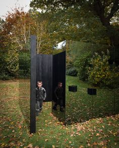 two young boys are standing in front of a mirrored sculpture on the grass, surrounded by trees and leaves