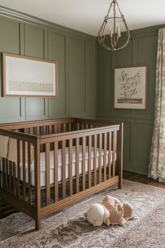 A gender-neutral nursery featuring a light wood crib, green paneled wall, boho-style rug, and beige couch with pillows, accented with framed wall art and greenery for a cozy, sophisticated feel. Brown And Green Nursery, Olive Green Nursery Neutral, Olive Green Baby Nursery, Green Nursery Colors, Evergreen Fog Nursery, Grey And Green Nursery, Sage Green Nursery Boy, Sage Green Baby Room, Nursery Ideas Green