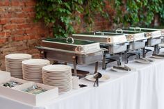 a buffet table set up with plates and silverware on it in front of a brick wall