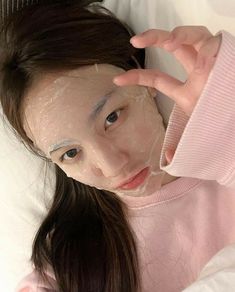 a woman laying on top of a bed holding her hand up to her face while wearing a pink sweater