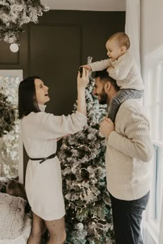 a man and woman are decorating a christmas tree with a baby on their shoulders