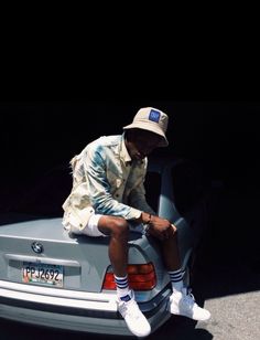 a man sitting on the trunk of a car wearing white sneakers and a hat with his hands in his pockets