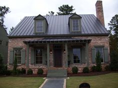 a brick house with a metal roof and two windows on the front porch, surrounded by grass and trees