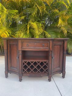 an old wooden cabinet with wine bottles on it's doors and bottom shelf, in front of some palm trees