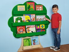 a young boy standing next to a green tree shaped book shelf with books on it