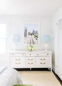 a white dresser with two lamps and a painting on the wall in front of it