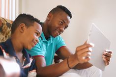 two young men sitting on a couch looking at something on a tablet computer while they both look at the screen