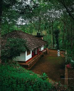 a small white house in the middle of some trees and dirt road with people walking around it