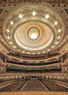 the inside of a large auditorium with wooden floors and ceiling lights on either side of the stage