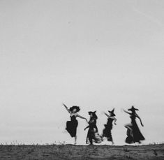 black and white photograph of four women in long dresses running across a field with one woman holding an umbrella