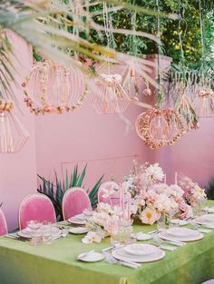 a green table topped with plates and pink chairs next to a wall covered in hanging chandeliers