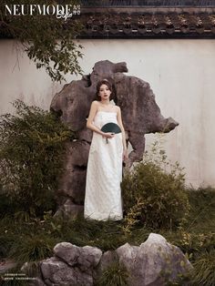 a woman in a white dress is holding a black plate and standing next to some rocks