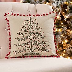 a christmas tree pillow on a white chair in front of a christmas tree with red pom - poms