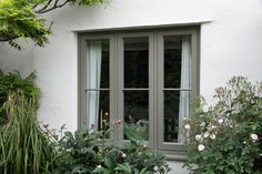 an open window on the side of a white building with green plants and flowers around it