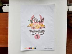 a cross - stitch picture of a deer with glasses and flowers on its head is hanging from a refrigerator