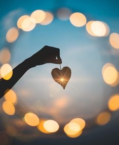 a person holding a heart shaped object in front of the sky with boket lights