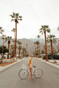 a woman standing next to a bike in the middle of a road with palm trees