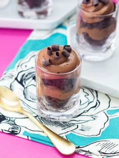 two glasses filled with dessert sitting on top of a pink and blue table cloth next to gold spoons