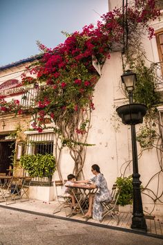 two people sitting at a table in front of a building with red flowers on it