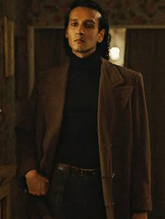 a man in a brown jacket and black turtle neck sweater standing next to a mirror