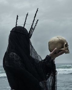 a woman holding a human skull in front of her face and wearing black veil on the beach