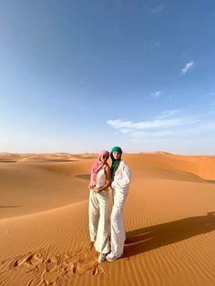 two people standing in the middle of a desert with sand dunes and blue sky behind them