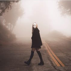 a woman walking down the middle of a road in front of trees and foggy sky