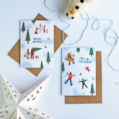 two christmas cards on a table next to a white ornament and paper snowflakes