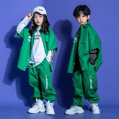 two children in green outfits standing next to each other against a blue wall, both wearing white sneakers