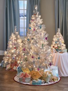 two white christmas trees with presents under them on a table in front of a window