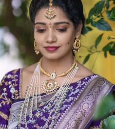 a woman in a purple and gold saree with jewelry on her neck, standing next to a tree