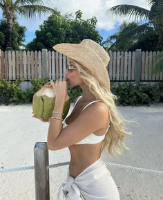 a woman wearing a straw hat drinking from a green coconut in front of a fence