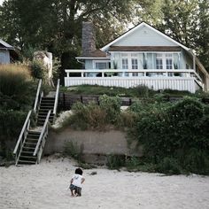 a young boy standing on top of a sandy beach next to a white house and trees