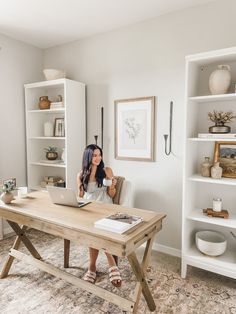 a woman standing in front of a desk with books on it and shelves behind her